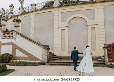 A bride and groom are walking down a stone staircase in front of a building. The bride is wearing a white dress and the groom is wearing a blue suit. The scene is romantic and intimate