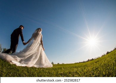 Bride And Groom Walking On The Grass And Holding Hands In A Park. Shot Is Taken With A Wide Angle Fish Eye Lens.