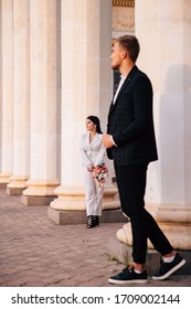 Bride And Groom In A White Pantsuit Posing On The Background Of Large White Columns