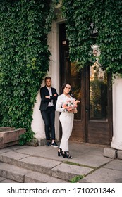 Bride And Groom In A White Pantsuit Posing Against The Backdrop Of A Building Overgrown With Ivy