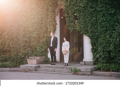 Bride And Groom In A White Pantsuit Posing Against The Backdrop Of A Building Overgrown With Ivy