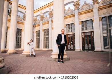 Bride And Groom In A White Pantsuit Posing On The Background Of Large White Columns