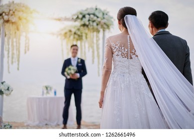 Bride with his father walking to the groom who waiting them with his flower bouquet. Seen on the beach in the evening. Happy wedding on the beach concept. 