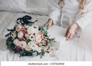 Bride Holding White Box For Morning Boudoir Before Wedding Ceremony.Modern Wedding Bouquet, Perfume Bottle, And Gift Box On White Bed.  Wedding Arrangements Or Bridal Shower