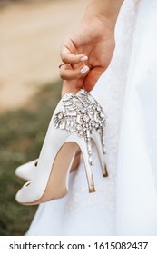 The bride holds in her hands white stained shoes with rhinestones. The girl is tired of walking in heels along the grass.
