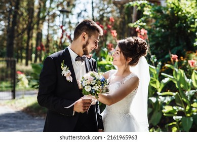 The bride looks at the groom. The groom in wedding suit and the bride in dress are standing and hugging in the Botanical green garden full of greenery. Wedding ceremony. - Shutterstock ID 2161965963