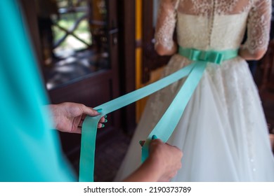 Bride In The Morning During Wedding Preparations. Bridesmaid Tying The Sash On The Bride's Dress