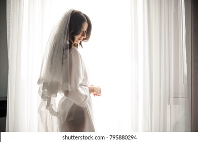 Bride Morning Preparation. Beautiful Bride In White Wedding Negligee And Veil On The Background Of Large Bedroom Window