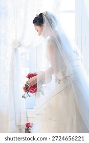 Bride prepares for wedding while holding bouquet near window in bright room with curtains