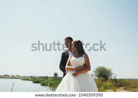 Bride in puffy dress is hugging with a groom in black suit near the lake. Wedding couple is standing on the hill outdoors. Romantic laughing summer love story. Azure blue water on the horizon.
