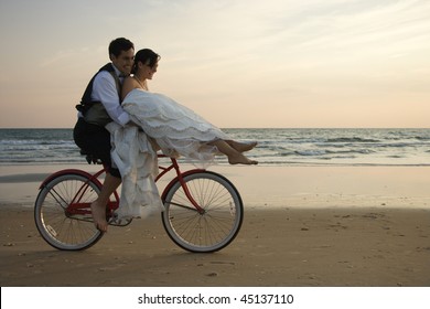 Bride rides the handle bars of a bicycle being driven by her groom on beach. Horizontal shot. - Powered by Shutterstock