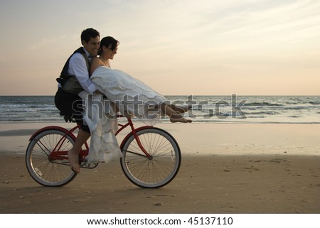 Bride rides the handle bars of a bicycle being driven by her groom on beach. Horizontal shot.