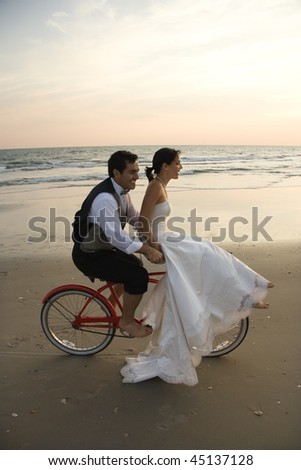 Bride rides the handle bars of a bicycle being driven by her groom on the beach. Vertical shot.