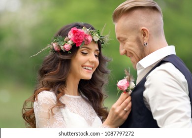 The Bride In Rustic Style With A Beautiful Wreath On The Head Of Flowers With A Bouquet In Hands  Standing In A Park In White Dress With Groom White Hair In White Shirt Suspenders And Black Waistcoat