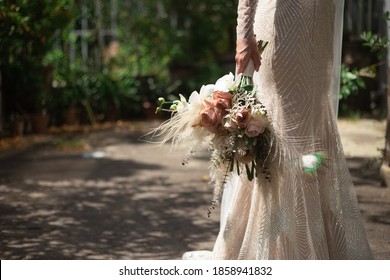 A Bride In A Shiny Wedding Dress Stands And Holds A Boho Style Wedding Bouquet In Her Hand.