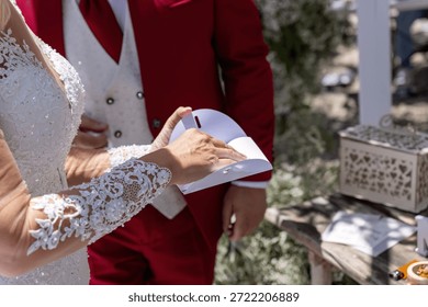 Bride signs wedding document while groom stands beside her in red suit during ceremony