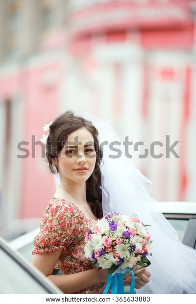 The bride in a simple retro dress with a floral\
pattern, already wearing a veil and holding  bouquet of a wedding,\
purple flowers, poses out of the car, vertical frame. Looking\
straight at the camera.