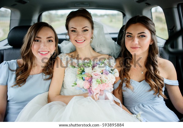 Bride sits between her bridesmaids in blue dress on\
back seat in the car