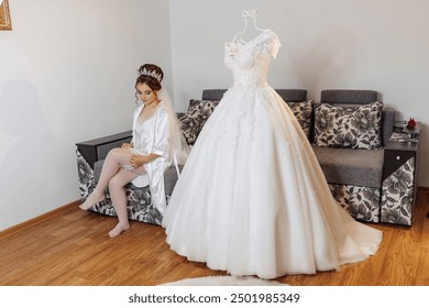 A bride is sitting on a couch in front of a wedding dress. The dress is white and has a long train - Powered by Shutterstock