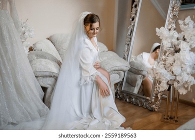 A bride is sitting on a couch in front of a mirror. She is wearing a white gown and a veil. The mirror reflects her image, and there is a vase of flowers in the background - Powered by Shutterstock