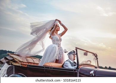 Bride Standing In Classic Convertible While Being Driven By Older Driver