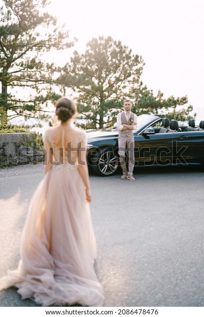 Bride stands across the street from groom\
leaning on the\
convertible