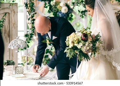 Bride stands behind groom while he signs wedding papers