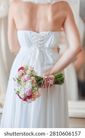 Bride stands with her back to the camera holding a pastel bouquet tied with ribbons, wearing a lace-up white wedding dress that conveys romantic, elegant, and intimate bridal preparation before the ceremony.