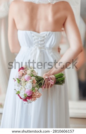 Bride stands with her back to the camera holding a pastel bouquet tied with ribbons, wearing a lace-up white wedding dress that conveys romantic, elegant, and intimate bridal preparation before the ceremony.