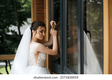 the bride stands at the window, lightly touching it with her hands. She has dark hair pulled back into an updo and a long white veil. Her reflection is visible in the glass of the window.