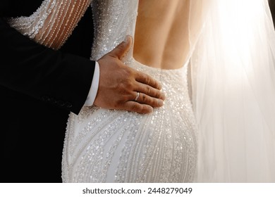 The bride wears a stunning white lace and pearl wedding dress with a long train. The groom is wearing a black tuxedo and has his hand on the brides waist. The couple is standing in a romantic pose. - Powered by Shutterstock