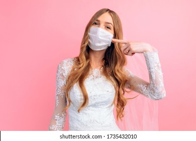 A bride in a wedding dress and a medical protective mask on her face points at the mask on a pink background. Wedding, quarantine, coronavirus - Powered by Shutterstock