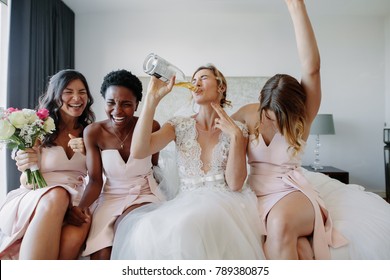 Bride in white dress drinking a bottle of wine while sitting on bed with bridesmaids. Bride and bridesmaids enjoying before wedding in hotel room.