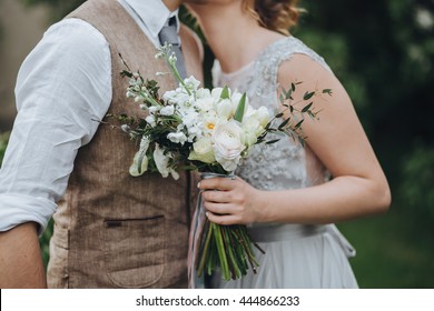 The bride in a white dress and groom in a waistcoat standing in the green garden, and holding a wedding bouquet