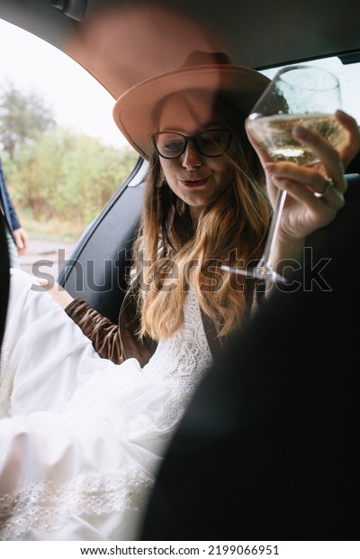 The bride in a white dress,\
leather jacket and hat sits in the back seat of the car, holding a\
bottle of champagne, a glass of champagne and\
smiling.