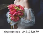 A bride in a white dress with puffy sleeves holds a wedding bouquet of bright pink peonies in her hands.