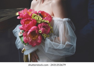 A bride in a white dress with puffy sleeves holds a wedding bouquet of bright pink peonies in her hands.: zdjęcie stockowe