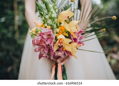 The bride in a white dress stands against a background of tropical plants and holds a bright bridal bouquet of flowers and greens with colored ribbons