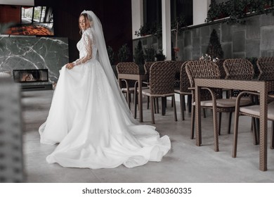 A bride in a white dress stands in front of a fireplace. She is wearing a veil and a long white gown. The scene is set in a restaurant with several chairs and tables - Powered by Shutterstock