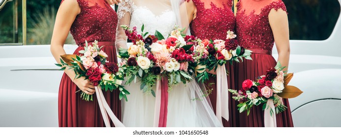 bride in white wedding dress and bridesmaids in red dresses are staying near the old white car with beige pink yellow and red flowers