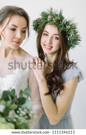 Bride in white wedding dress standing with her smiling bridesmaid in pine wreath and blue dress