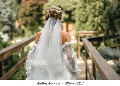 Bride In A White Wedding Dress And Veil With A Beautiful Hairstyle Back View. Cute Blonde Bride Walking On A Wooden Bridge On A Background Of Trees