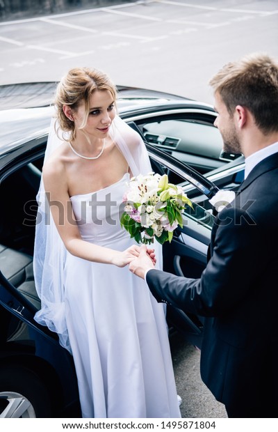 bridegroom in suit giving hand to attractive and\
smiling bride with\
bouquet