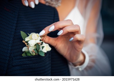 Bride's hand touches the buttonhole on the groom's jacket, close-up. Stylish suit, boutonniere of fresh flowers, woman's hand with a manicure.