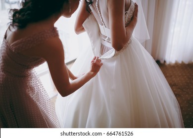 Bridesmaid helping slender bride lacing her wedding white dress, buttoning on delicate lace pattern with fluffy skirt on waist. Morning bridal preparation details newlyweds. Wedding day moments, wear. - Powered by Shutterstock