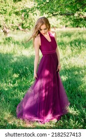 Bridesmaid Stands In The Summer Garden And Demonstrates A Festive Dress Of Purple Color, Posing Looking Down. Professional Makeup And Hair Styling, High Quality Photo