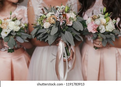 Bridesmaids and bride holding modern wedding bouquets of pink roses and green eucalyptus with pink ribbons. Stylish Contemporary bouquets on soft fabric. Wedding arrangements