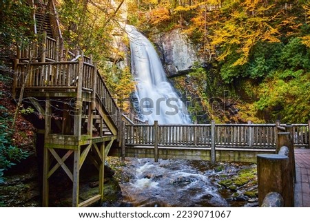 Bridge boardwalk crosses river in front of huge waterfall over cliffs surrounded by fall foliage