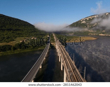 A bridge connects two sides of a river as fog rises in the morning. Trees and hills surround the area. Trains travel on the rails beside the water.