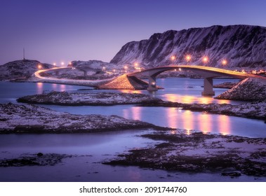 Bridge With Illumination, Snow Covered Mountains, Village And Violet Sky With Reflection In Water. Night Landscape With Bridge, Snowy Rocks, City Lights, Sea. Winter In Lofoten Islands, Norway. Road